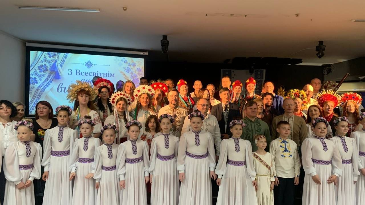 Групове фото учасників святкового заходу до Всесвітнього дня вишиванки на сцені. На передньому плані стоять дівчата в однакових білих сценічних сукнях та віночках, а за ними — дорослі у різноманітних вишиванках та традиційних українських головних уборах на тлі екрана з вітальним написом.