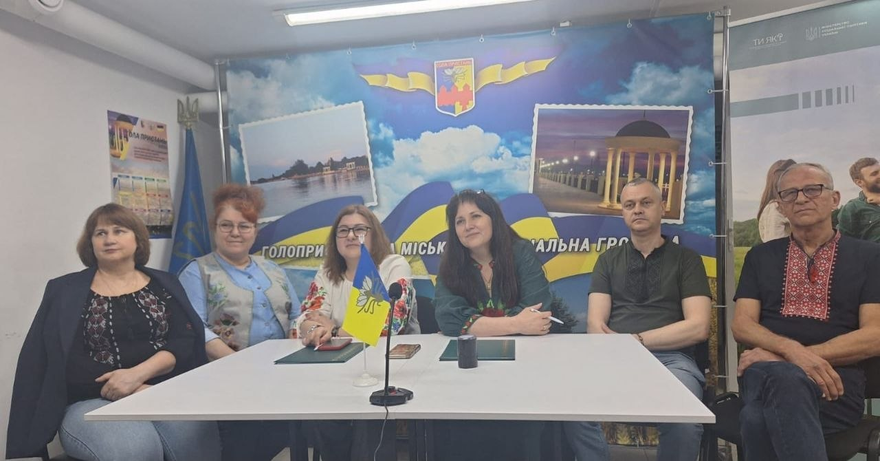 Групове фото шістьох людей у приміщенні, які сидять за білим столом на тлі великого синьо-блакитного банера з написом «Голопристанська міська територіальна громада». На столі встановлено маленький прапорець Голої Пристані, а декілька присутніх вбрані у сорочки з вишивкою.