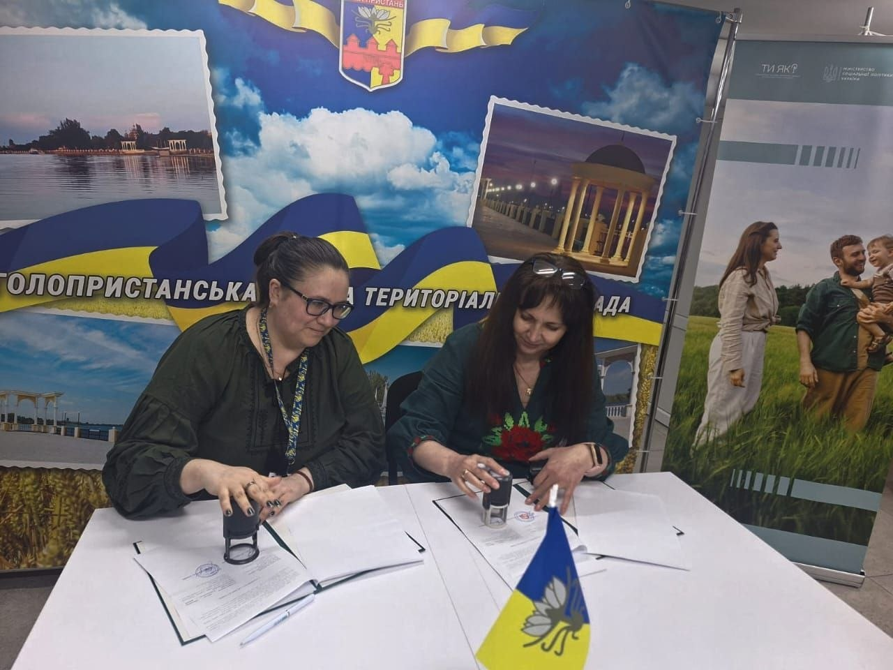 Дві жінки в темних вишиванках сидять за білим столом і ставлять печатки на документи. Вони позують на тлі великого синьо-блакитного банера з написом «Голопристанська міська територіальна громада», а на столі перед ними встановлено маленький прапорець міста Гола Пристань.