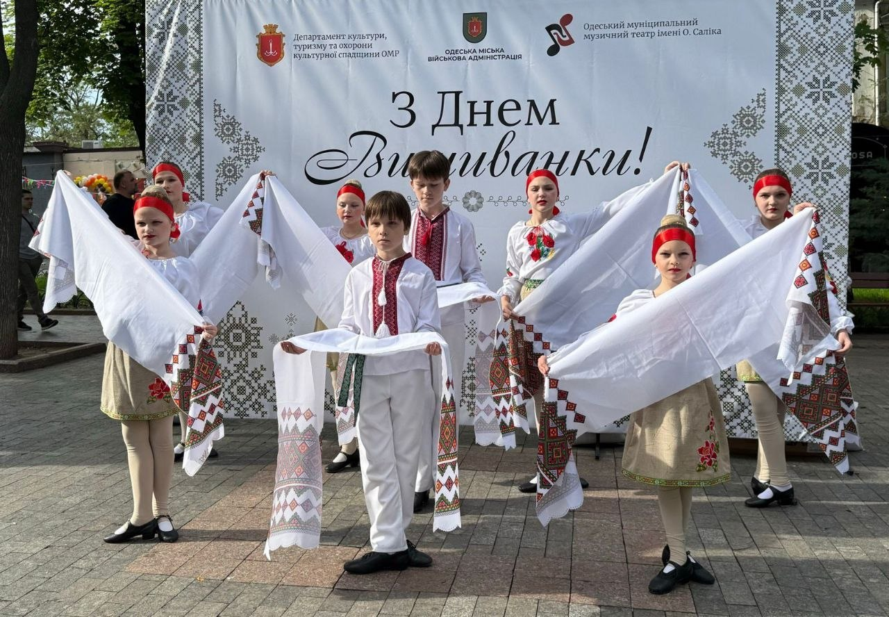 Виступ дитячого танцювального колективу на вулиці на тлі великого світлого банера з написом «З Днем Вишиванки!». Діти вбрані в традиційні українські костюми, червоні пов'язки на голові та тримають у руках довгі вишиті рушники.