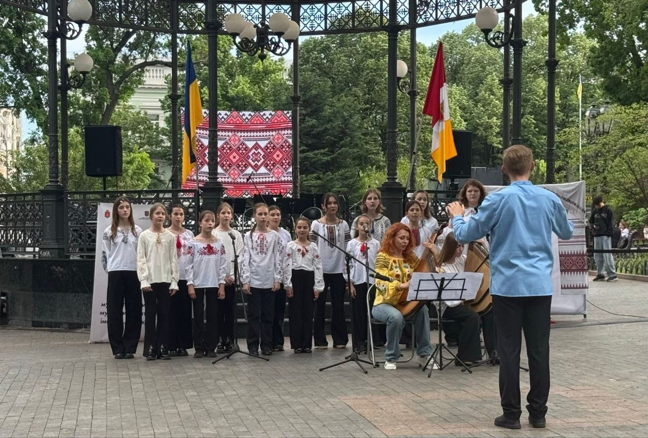 Виступ дитячого хору в білих вишиванках просто неба на тлі великої кованої альтанки. Перед хором стоїть диригент спиною до камери, поруч жінка акомпанує на бандурі, а на задньому плані розміщено екран із традиційним червоно-білим орнаментом та прапори.