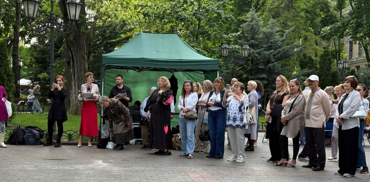Група людей у літньому одязі та вишиванках спостерігає за заходом просто неба в парку. Вони стоять на брукованій майданчику біля великого зеленого намету, навколо якого ростуть високі дерева та встановлені декоративні ліхтарі.