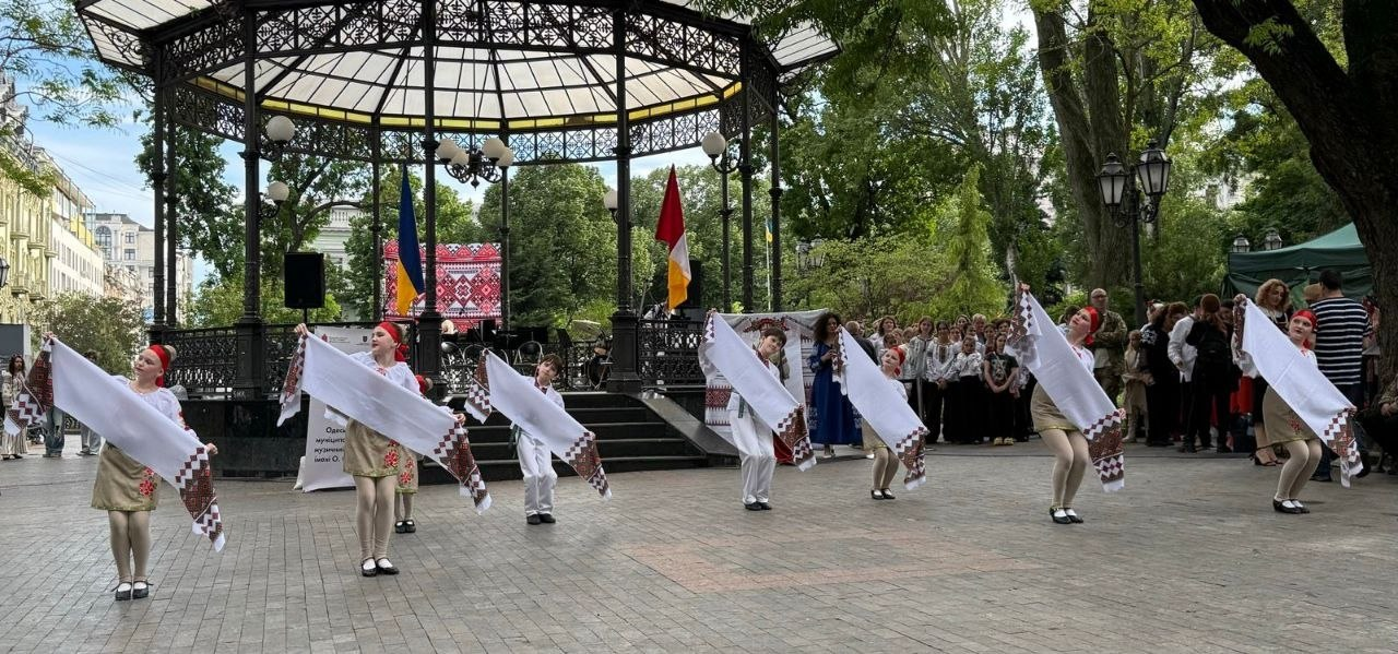 Виступ дитячого танцювального колективу в традиційних українських костюмах просто неба на брукованому майданчику. Діти тримають у руках довгі вишиті рушники на тлі великої критої кованої альтанки та глядачів у парку.