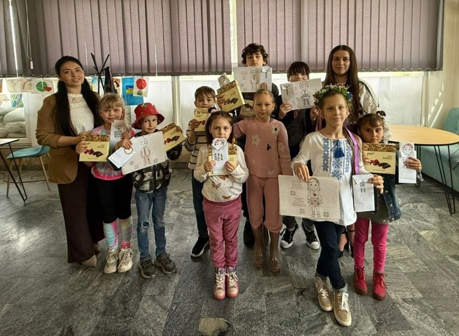 Групове фото дітей та двох дорослих жінок у приміщенні з великими вікнами та жалюзі. Діти тримають у руках коробки цукерок, малюнки та паперові вироби, а одна дівчинка на передньому плані вбрана у вишиванку та віночок зі стрічками.