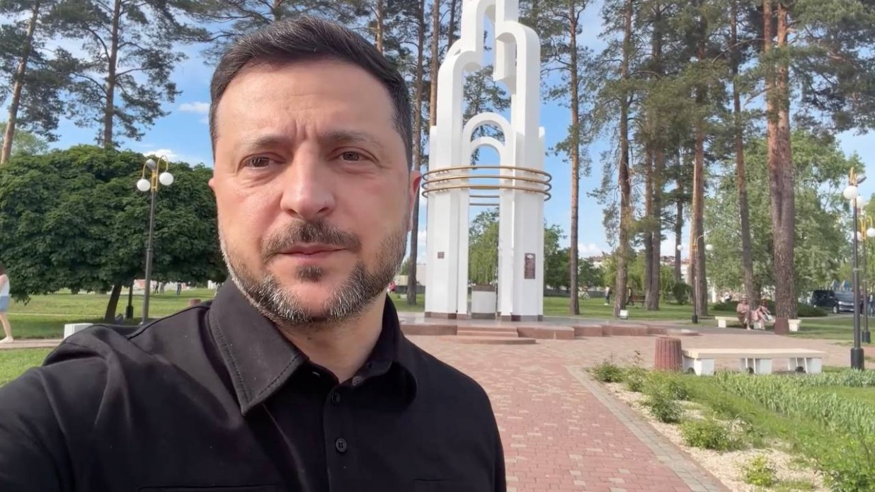 Чоловік у чорній сорочці робить селфі-знімок просто неба на тлі парку з високими соснами та брукованою доріжкою. На задньому плані по центру розташована велика біла архітектурна споруда-монумент у вигляді арок із золотистими елементами.