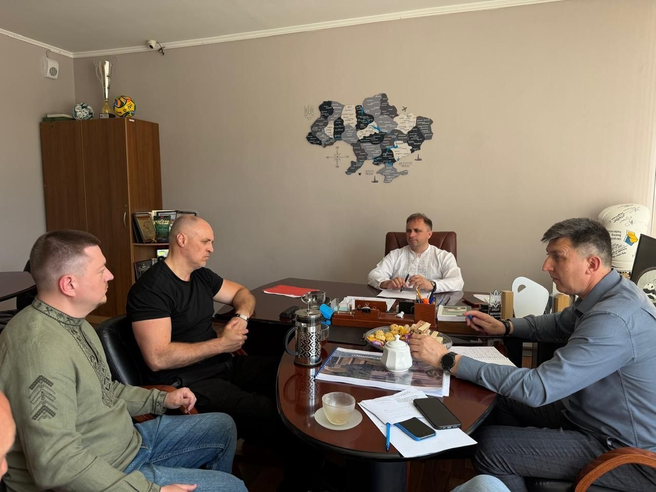 Група чоловіків проводить робочу зустріч за великим столом у кабінеті. На стіні позаду них розміщено декоративну мапу України, а на столі лежать документи і канцелярія.