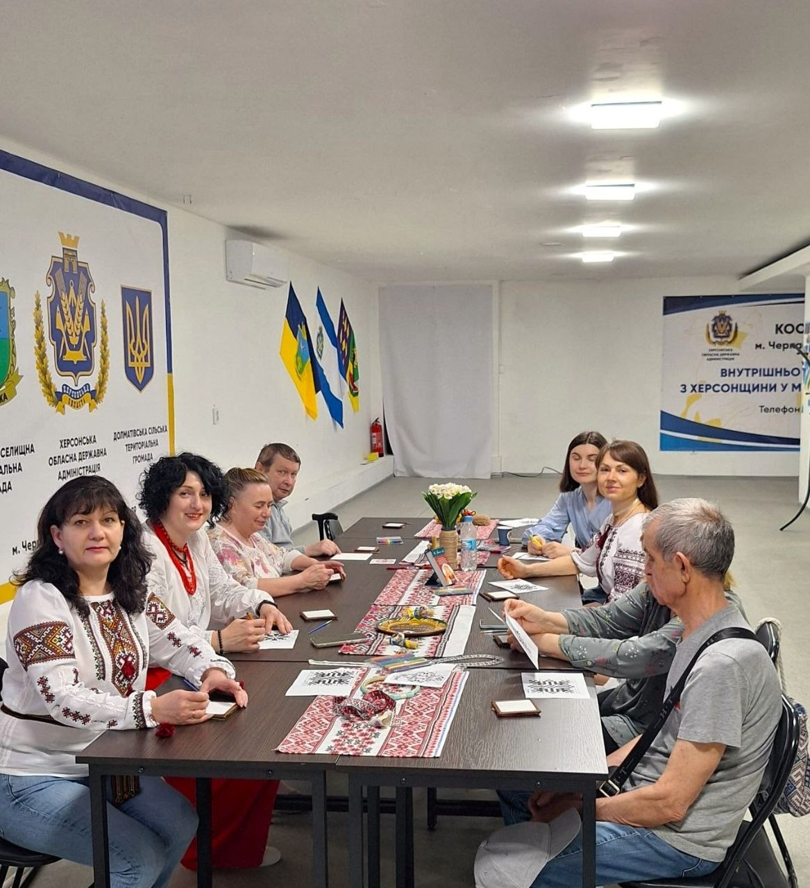 Група людей сидить за довгим столом у світлому кабінеті під час офіційного заходу чи майстер-класу. Кілька учасниць одягнені в українські вишиванки, на столі лежать папери та декоративні елементи, а на стінах розміщено прапори, герби та інформаційні банери.