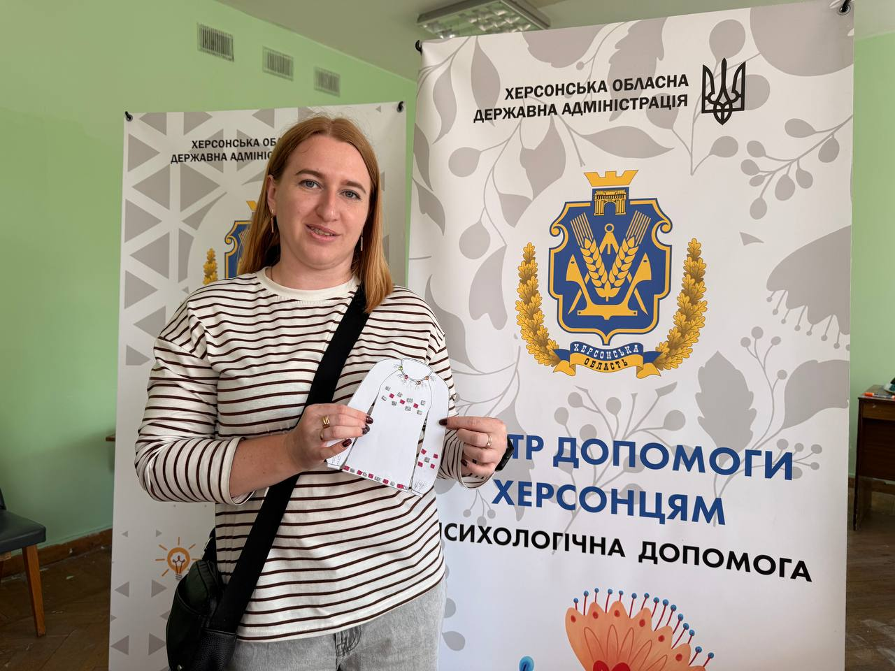 Жінка в смугастій кофті стоїть у кабінеті та тримає в руках невеличку білу паперову саморобку у формі сорочки з вишивкою. Вона позує на тлі банерів Херсонської обласної державної адміністрації з написами про психологічну допомогу та гербом області.