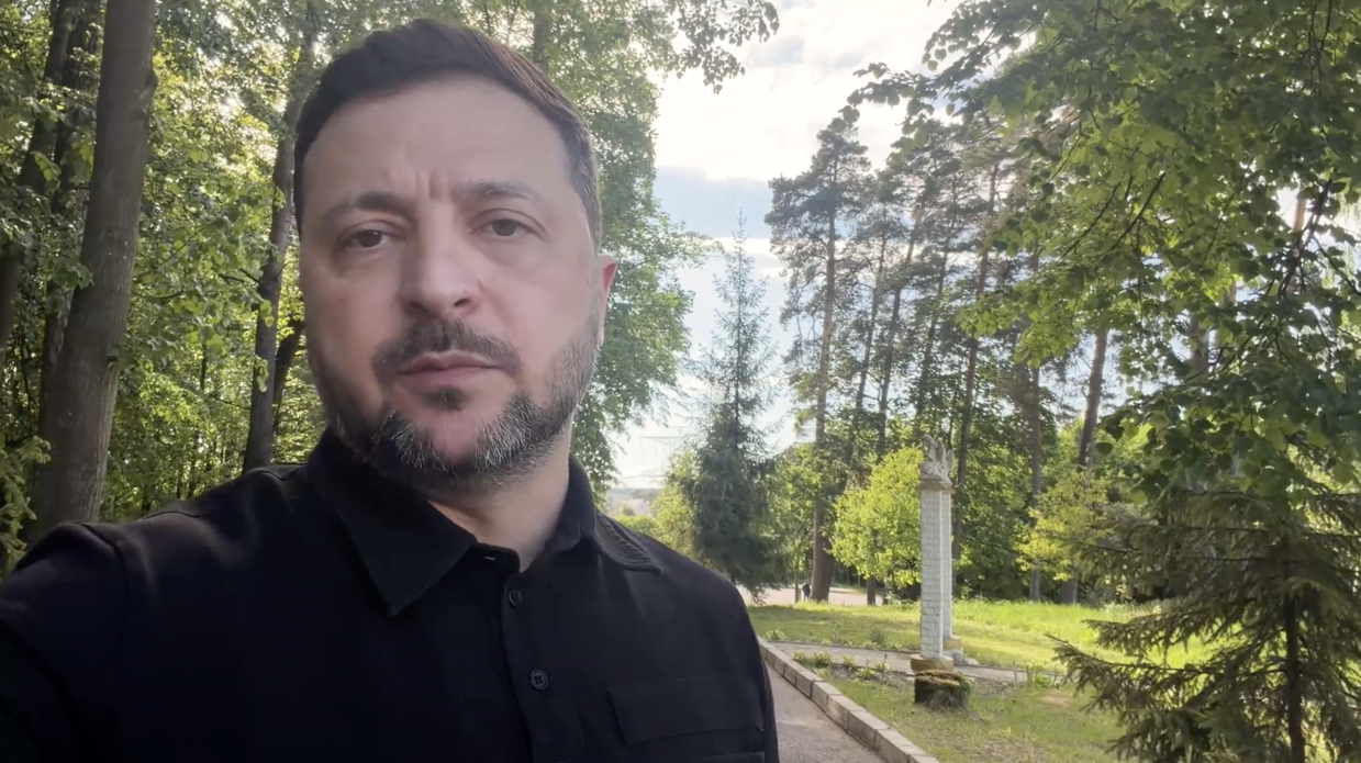 Чоловік у чорній сорочці знятий великим планом на селфі-камеру під час звернення на вулиці. Він стоїть на тлі зеленого парку з високими хвойними та листяними деревами під світлим небом.