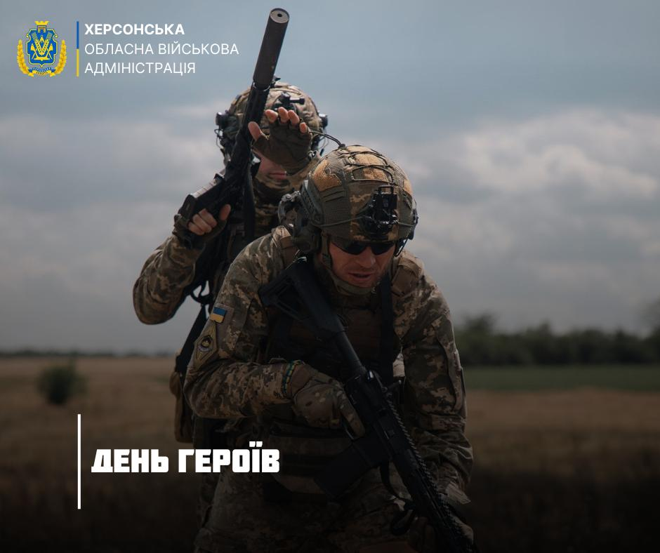 Українські військові у камуфляжній формі та захисному спорядженні під час виконання бойового завдання у полі. На зображенні присутній логотип Херсонської обласної військової адміністрації та напис «День Героїв».