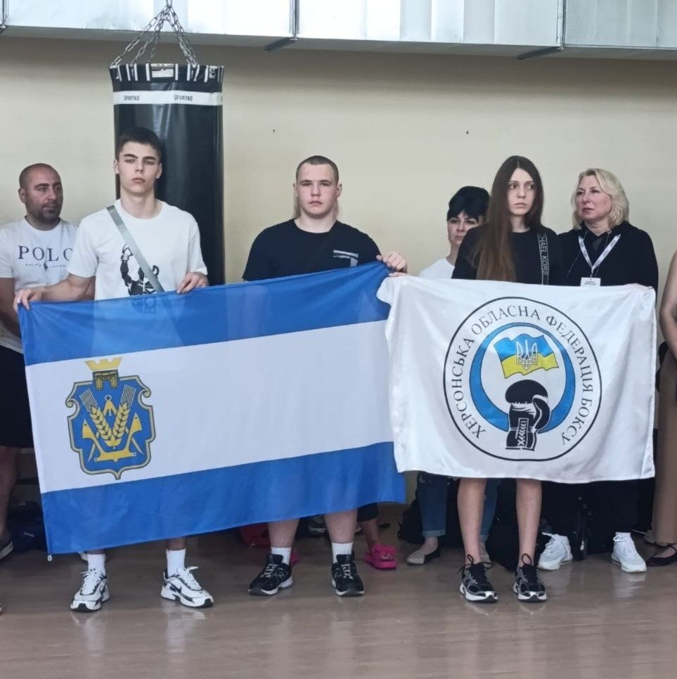 Група людей у спортивному залі тримає два прапори: офіційний прапор Херсонської області та білий прапор із логотипом «Херсонська обласна федерація боксу». На задньому плані видно боксерську грушу.