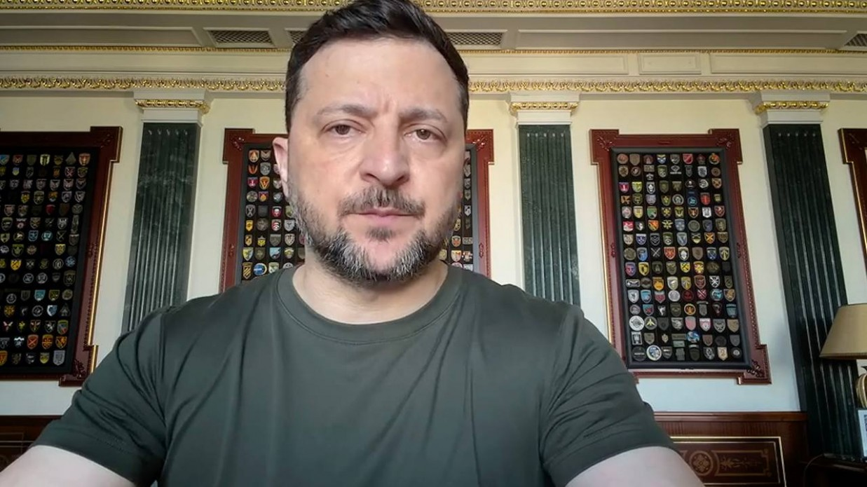 Чоловік із короткою бородою та в темно-зеленій футболці сидить у кабінеті, на стінах якого позаду нього висять декоративні стенди з шевронами та військовими емблемами.