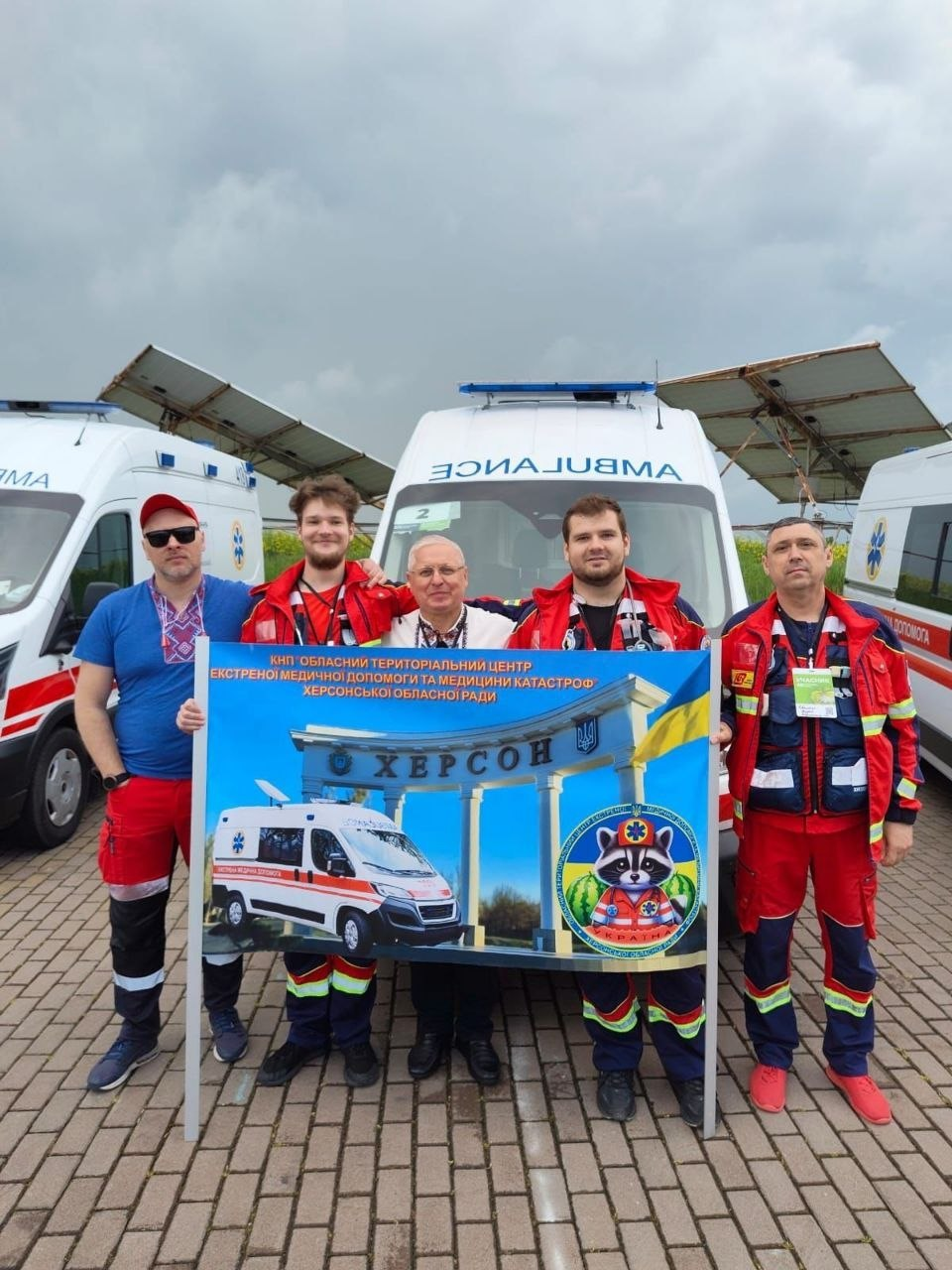 П'ятеро чоловіків у медичній формі стоять на тлі автомобілів швидкої допомоги та тримають великий інформаційний банер із написом «Херсон».