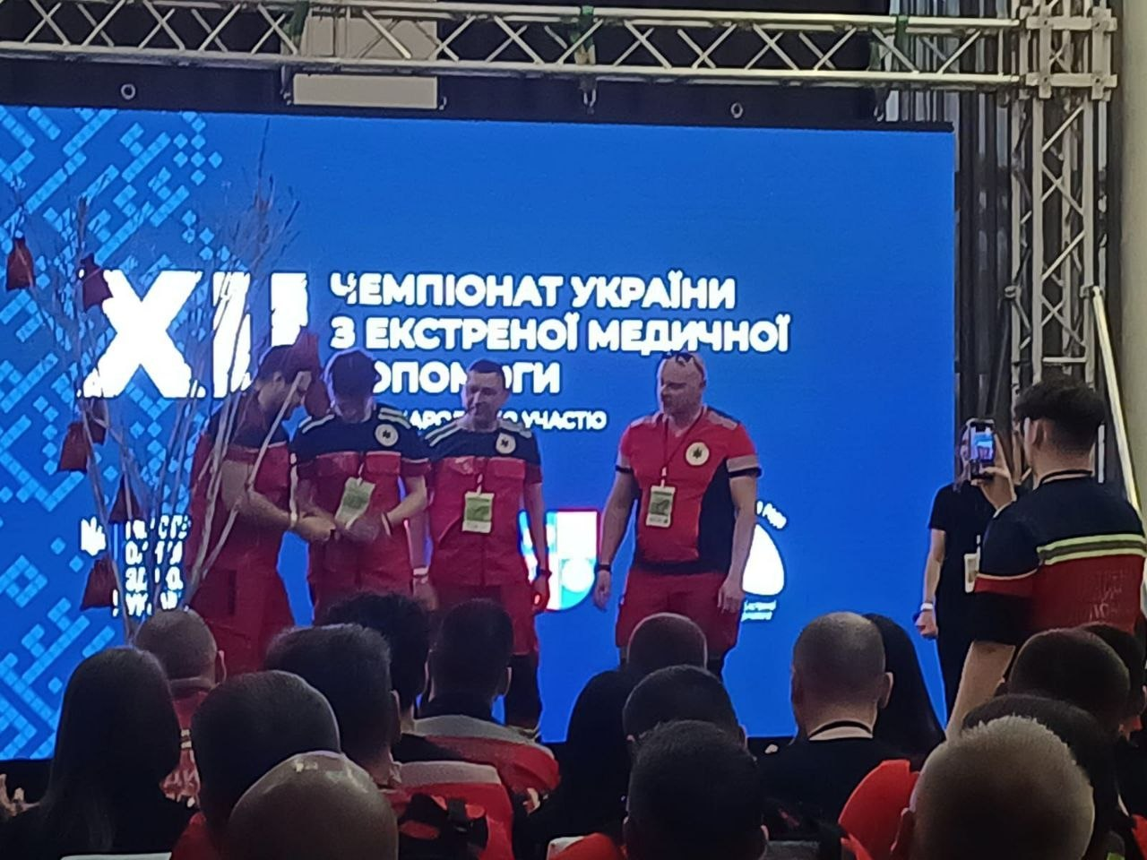 Група медиків у червоній формі стоїть на сцені перед великим синім екраном із написом «ХІ Чемпіонат України з екстреної медичної допомоги» під час офіційного заходу перед глядачами.