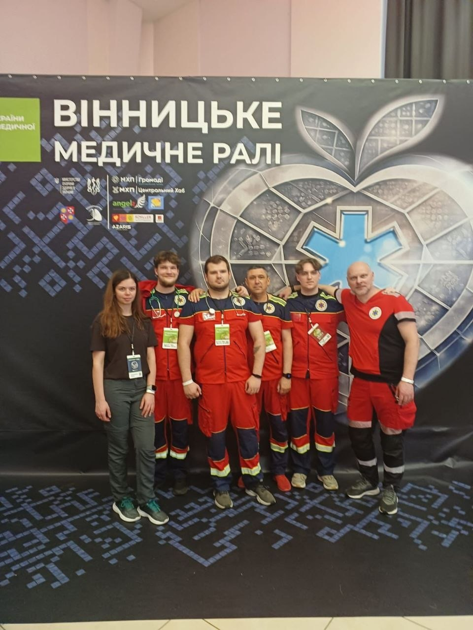 Група людей з шести осіб стоїть перед великим темним банером з написом «Вінницьке медичне ралі». Перша ліворуч дівчина одягнена у повсякденний одяг, а п'ятеро чоловіків праворуч від неї — у червоно-синю медичну уніформу екстреної допомоги.