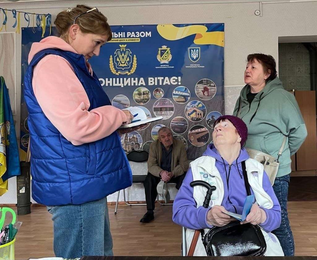 Жінка-волонтер у синьому жилеті робить записи в журналі, спілкуючись із літньою жінкою з палицею та іншими відвідувачами у приміщенні гуманітарного центру на тлі патріотичного банера.