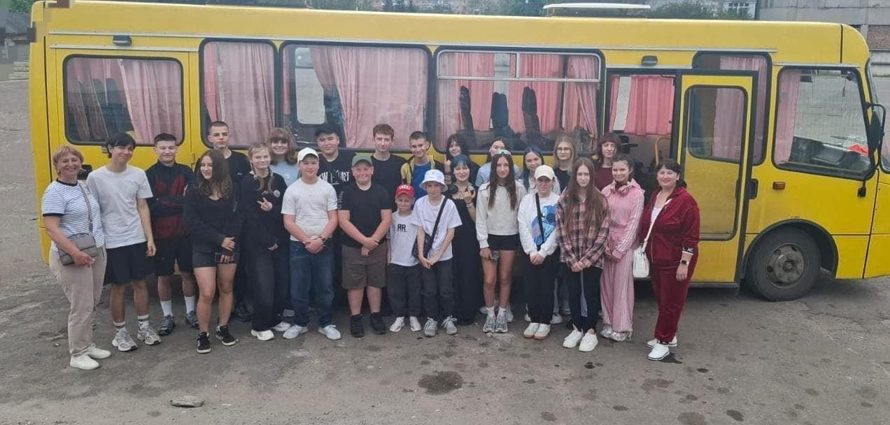 Група підлітків та дорослих стоїть на асфальтованому майданчику поруч із великим жовтим автобусом, позичуючи для спільного групового фото перед поїздкою.