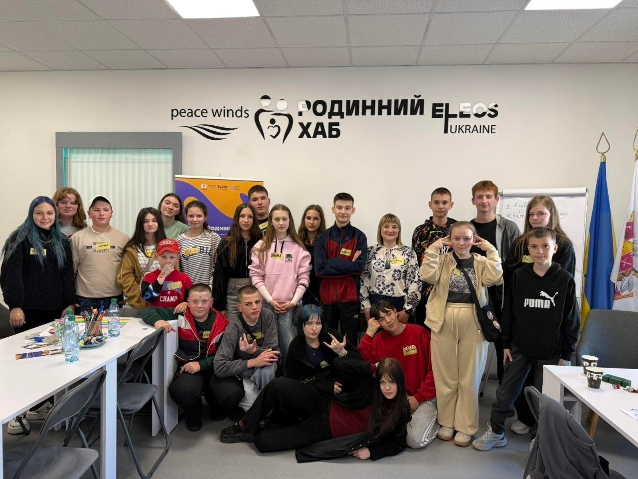 Група дітей та підлітків позує для спільного фото у світлому кабінеті на тлі стіни з логотипами благодійних організацій та написом «Родинний хаб».