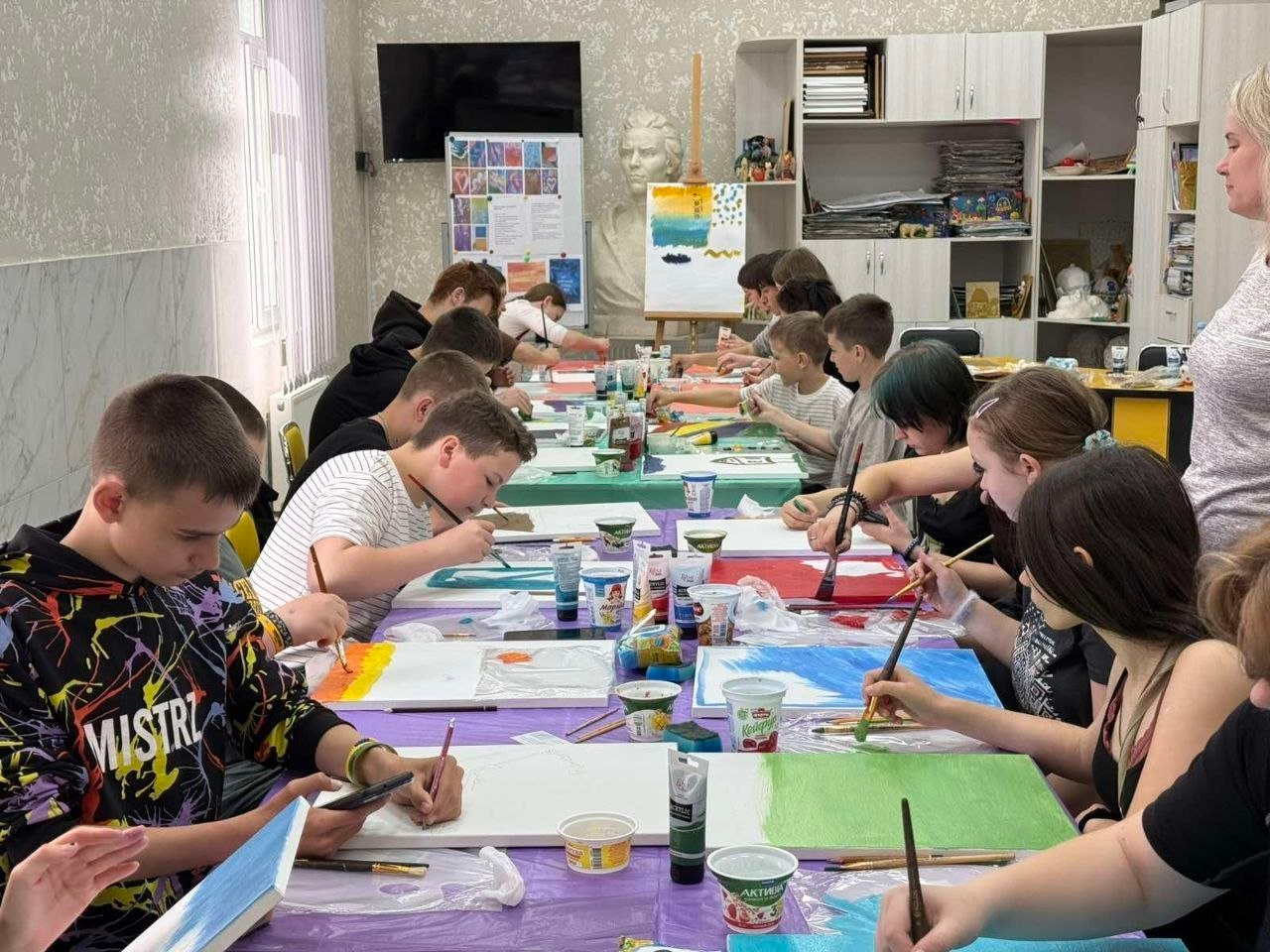 Група дітей та підлітків сидить за довгим столом у світлому навчальному кабінеті чи художній студії та малює пензлями на полотнах під час творчого майстер-класу.