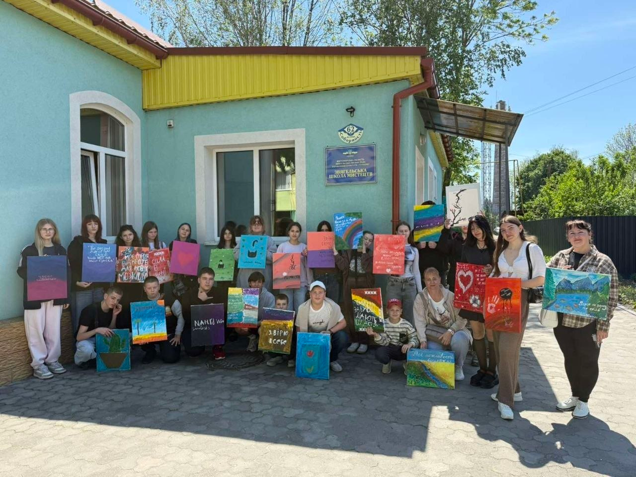 Велика група дітей, підлітків та дорослих стоїть на подвір'ї біля будівлі школи мистецтв, тримаючи в руках свої власні малюнки на полотнах під час спільного фото на вулиці в сонячний день.