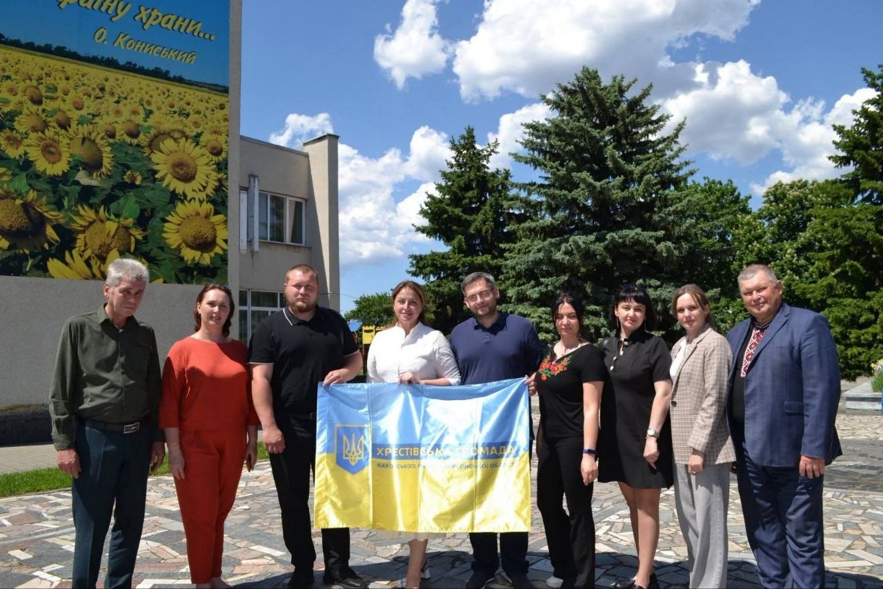Група людей стоїть на вулиці в сонячний день на тлі будівлі та хвойних дерев, розгорнувши синьо-жовтий прапор Хрестівської громади Херсонської області. Ліворуч на стіні будівлі розміщено великий патріотичний плакат із зображенням соняшникового поля та рядками поезії Олександра Кониського.