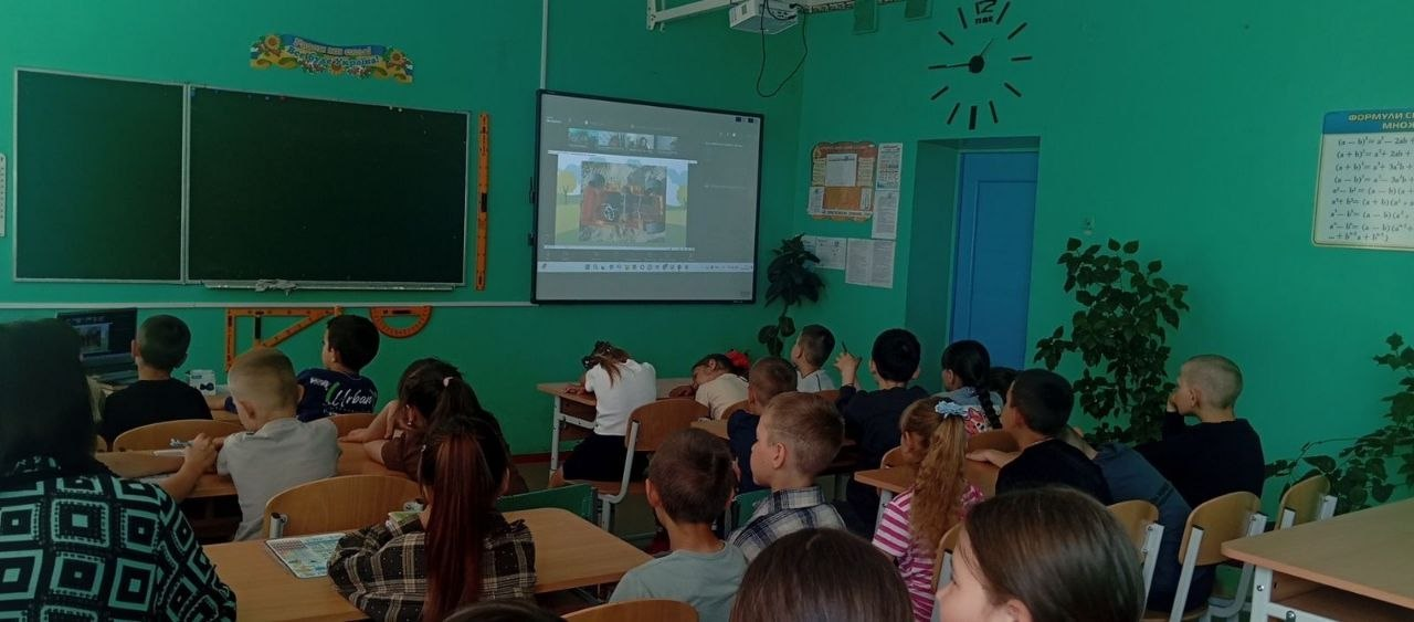 Учні молодших класів сидять за партами у шкільному кабінеті з зеленими стінами та дивляться на великий інтерактивний екран, де демонструється відеоурок під час занять.