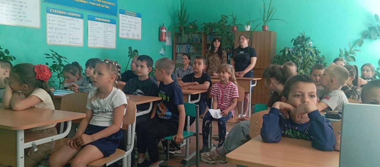 Група дітей та дорослих під час заняття чи заходу в шкільному кабінеті математики з зеленими стінами, де на задньому плані стоять стелажі з книгами та кімнатними рослинами, а на стіні висять навчальні плакати з формулами.