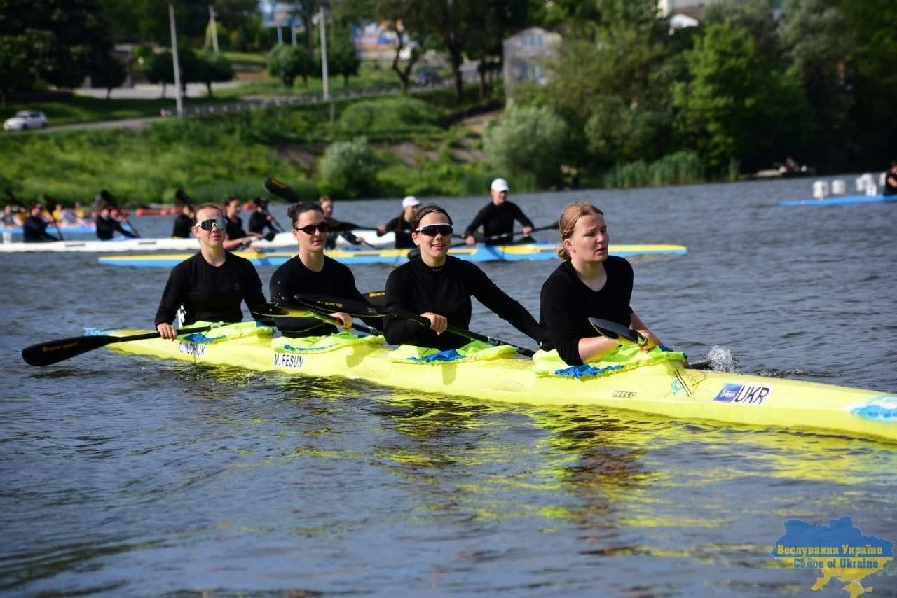 Чотири спортсменки в чорному одязі веслують на довгій яскраво-жовтій байдарці-четвірці під час змагань або тренувань на річці. На задньому плані видно інших веслярів у човнах та зелений берег, а в нижньому кутку розміщено логотип «Веслування України / Canoe of Ukraine».