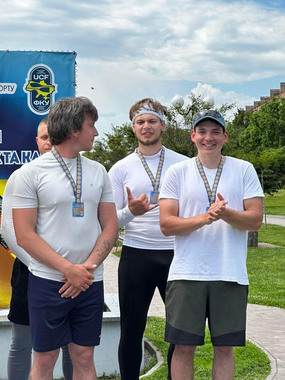 Три спортсмени у спортивному одязі з медалями на шиях стоять поруч на вулиці біля банера з логотипом Федерації каное України під час церемонії нагородження.
