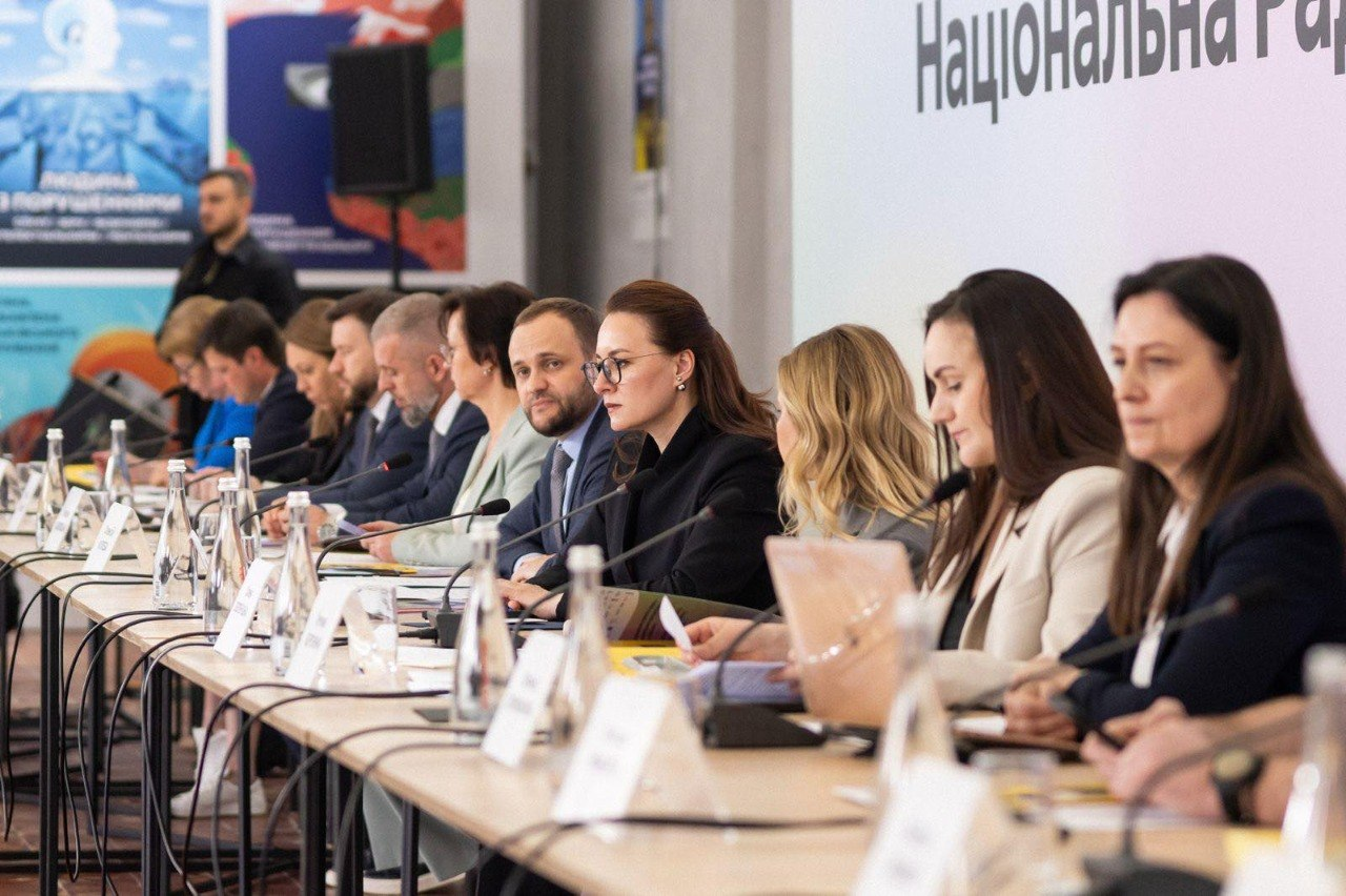 Група чоловіків та жінок у діловому одязі сидить у ряд за довгим столом із мікрофонами, пляшками води та паперами під час офіційного засідання в конференц-залі.