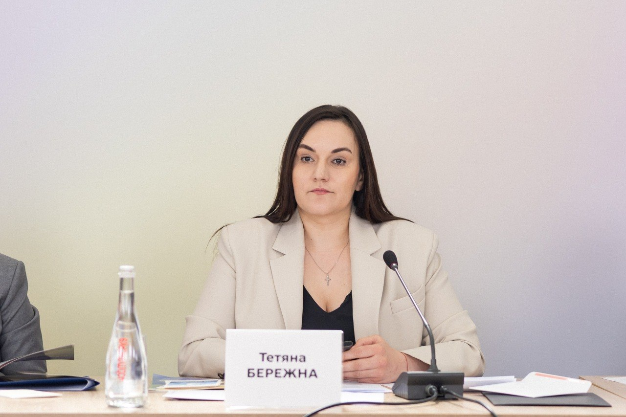 Жінка у світлому піджаку сидить за столом під час офіційного заходу перед настільним мікрофоном, документами та табличкою з ім'ям на нейтральному тлі.