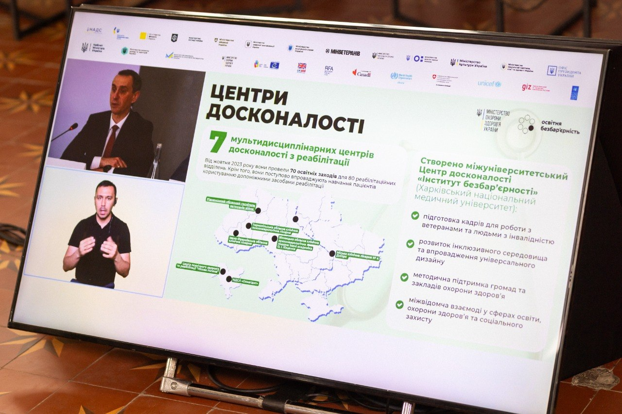 Скриншот екрана з презентацією про «Центри досконалості» та реабілітацію, де на слайді зображено карту України з позначками, текстові блоки та логотипи, а ліворуч відображаються вікна з доповідачем та сурдоперекладачем.