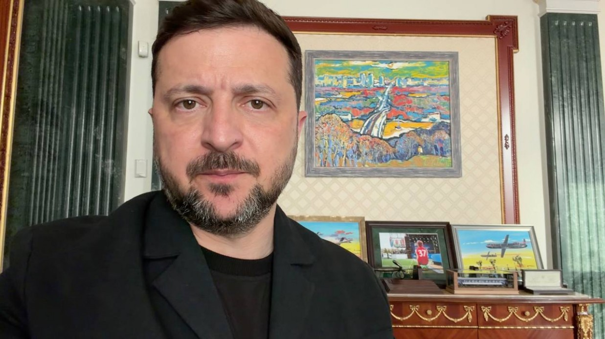 Чоловік із короткою бордою та в чорному піджаку зафіксований на передньому плані під час відеозвернення, а на задньому плані видно інтер'єр кабінету з картинами на стіні та комоді, серед яких зображення пейзажу та літака.