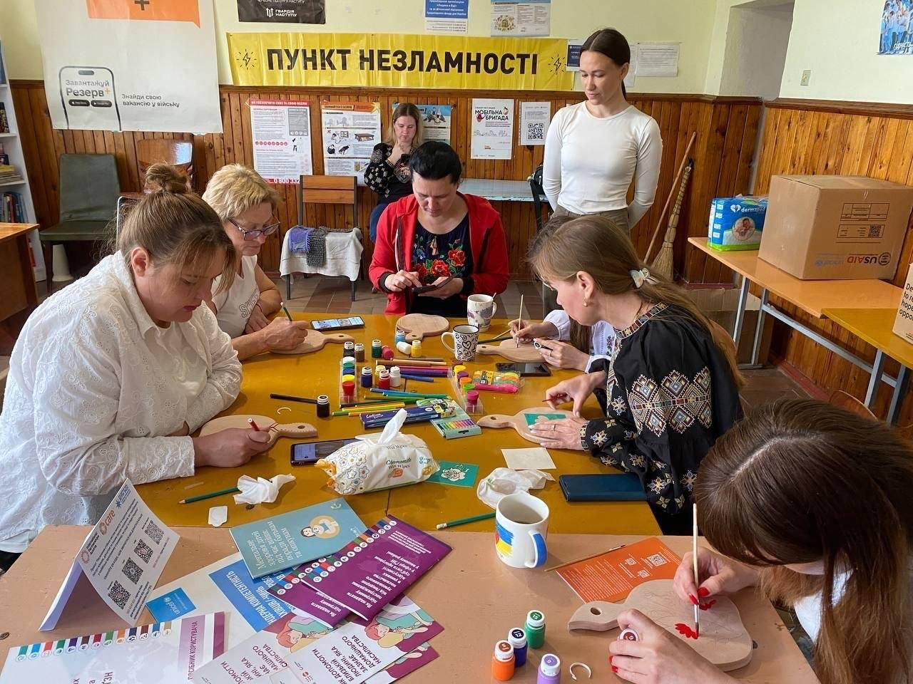 Група жінок сидить за столом у «Пункті незламності» та розфарбовує дерев'яні дощечки. На столі розкладені пензлі, фарби, олівці та кольорові буклети. Дві жінки стоять поруч, а на стіні позаду висять інформаційні плакати.