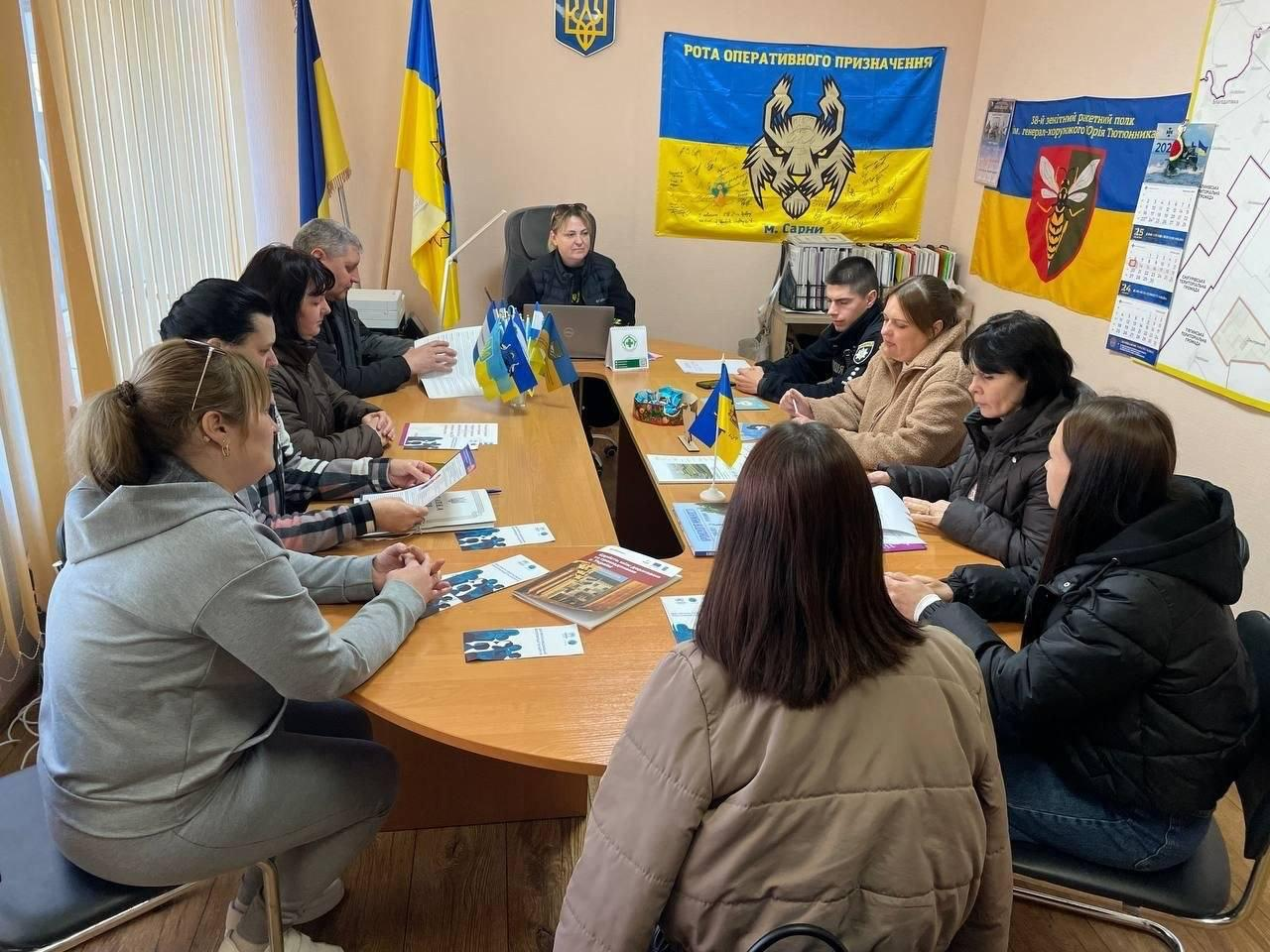 Група людей сидить навколо великого овального столу під час робочої зустрічі чи наради в кабінеті. На столі розкладені документи, буклети та маленькі прапорці України. На стінах кабінету висять державні прапори, великий синьо-жовтий стяг із символікою роти оперативного призначення та настінний календар.