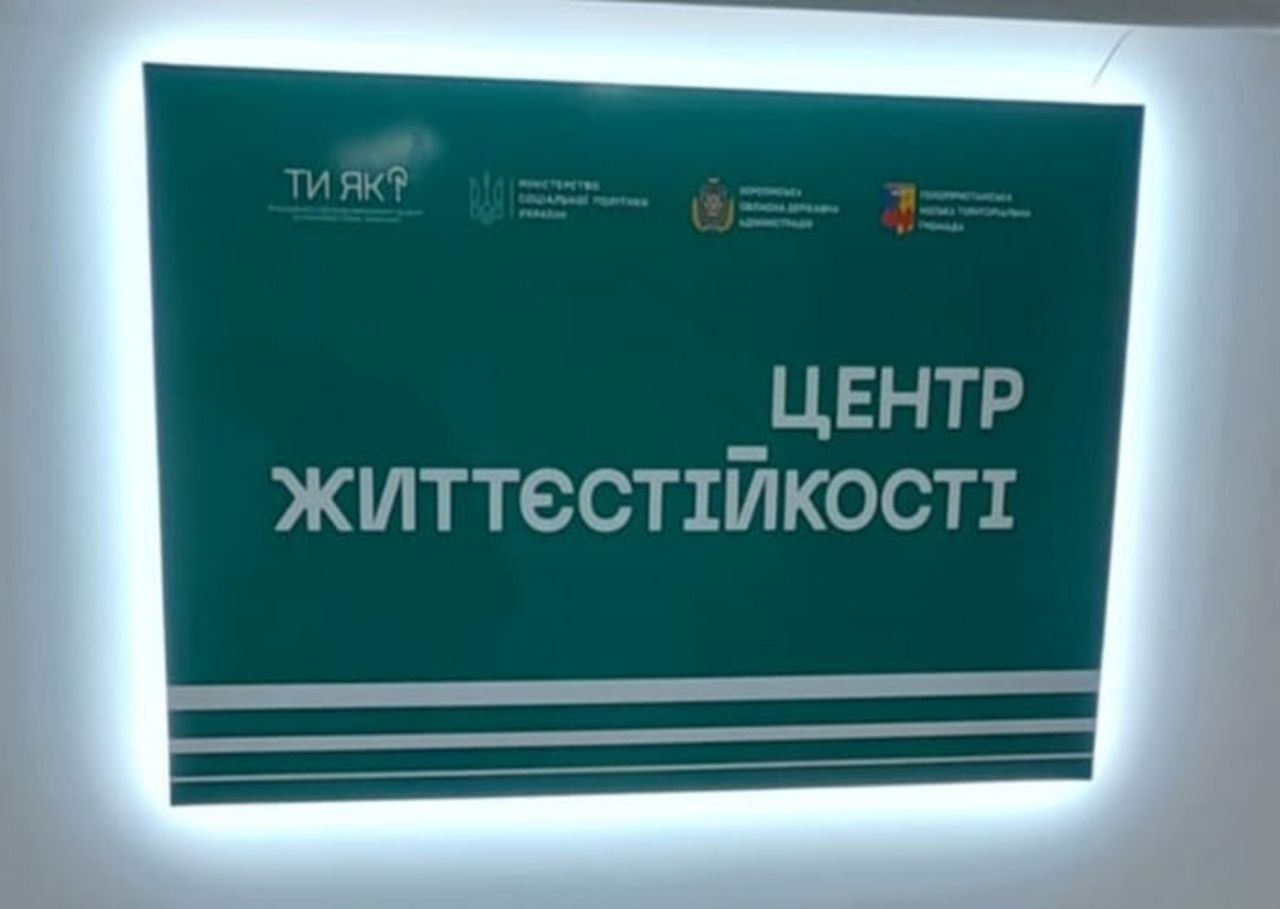 Інформаційна вивіска «Центр Життєстійкості» зеленого кольору із внутрішнім підсвічуванням на білій стіні. У верхній частині вивіски розміщено логотипи програми «Ти як?», Міністерства соціальної політики України, Херсонської ОДА та Великоолександрівської громади.
