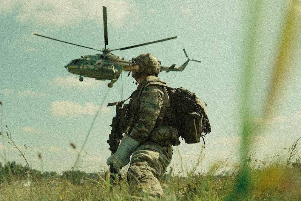 Військовослужбовець у камуфляжній екіпіровці з каскою та рюкзаком стоїть у полі серед трави й дивиться на військовий гелікоптер, що летить у небі.