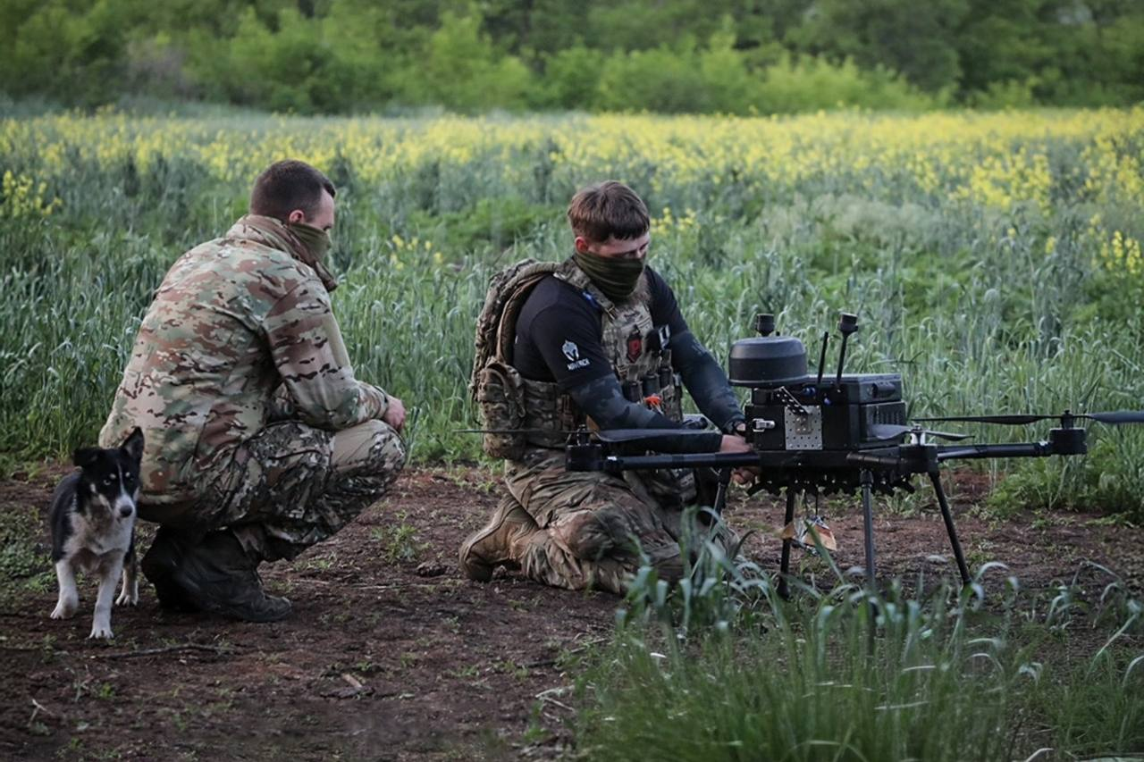 Два військовослужбовці з прикритими обличчями налаштовують великий безпілотник мультироторного типу на землі біля поля, а поруч із ними стоїть невеликий собака.