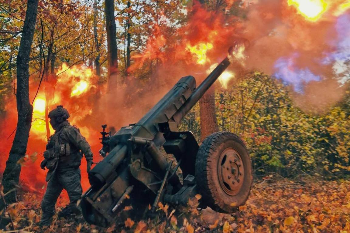 Постріл військової артилерійської гармати у лісистій місцевості восени: з дула виривається велике полум'я та дим, а поруч у захисному спорядженні стоїть військовослужбовець.