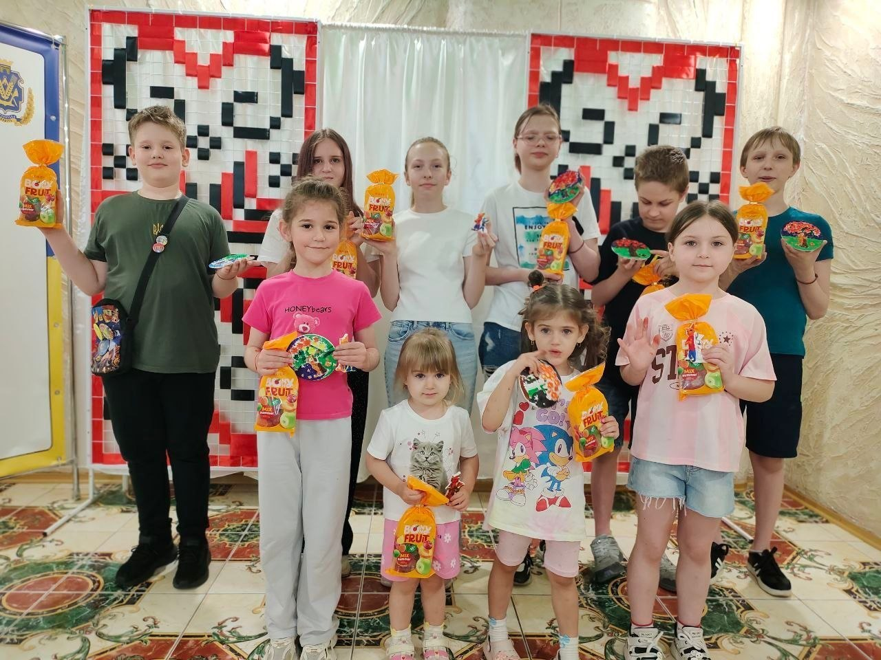 Група дітей різного віку стоїть у приміщенні та тримає в руках солодощі в помаранчевих пакунках і сувеніри. На тлі стіни розміщено декоративні полотна з традиційним українським червоно-чорним орнаментом.