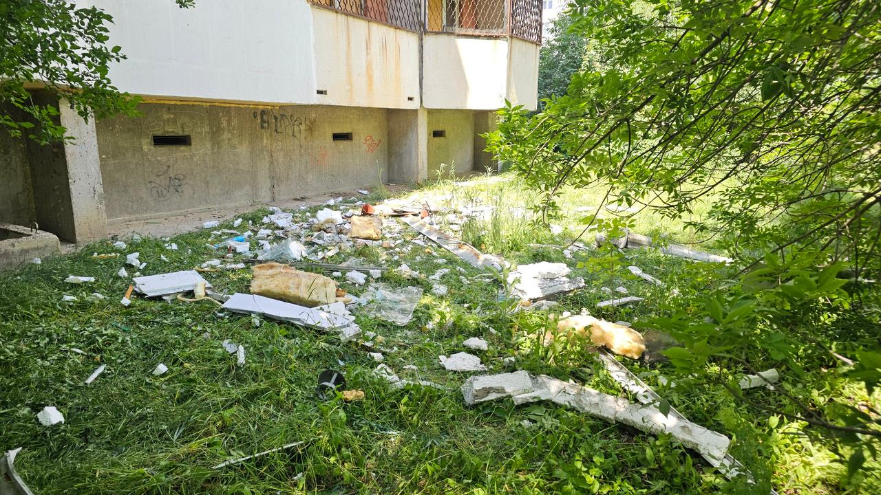 Засмічена та всіяна будівельними уламками зелена галявина біля підніжжя житлового будинку з відкритим цокольним поверхом в оточенні густих дерев.