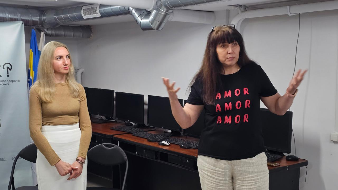 Дві жінки виступають перед аудиторією в приміщенні з комп'ютерними робочими місцями. Спікерка праворуч у чорній футболці з написом «AMOR» активно жестикулює руками під час промови, а дівчина ліворуч у бежевій кофті та білій спідниці слухає її.