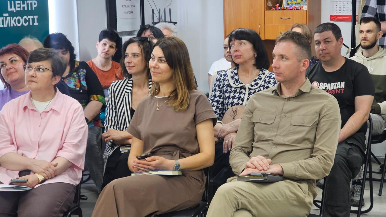 Група людей сидить у залі та уважно слухає виступ. На передньому плані видно жінку в коричневій сукні та чоловіка в сорочці кольору хакі з блокнотами в руках. Ліворуч на задньому плані частково видно вивіску «Центр життєстійкості».