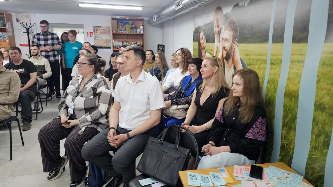 Група людей сидить у залі під час лекції. На стіні праворуч розміщено велике фотошпалерне зображення родини на тлі зеленого поля, а на передньому плані на столі розкладено інформаційні буклети.