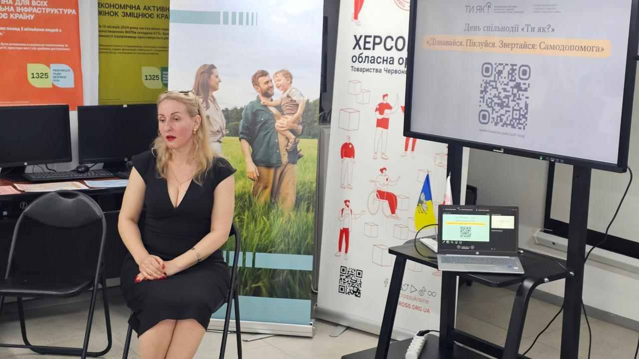 Жінка у чорній сукні сидить на стільці в залі для презентацій. Праворуч від неї встановлено великий екран і ноутбук із презентацією «День спільнодії