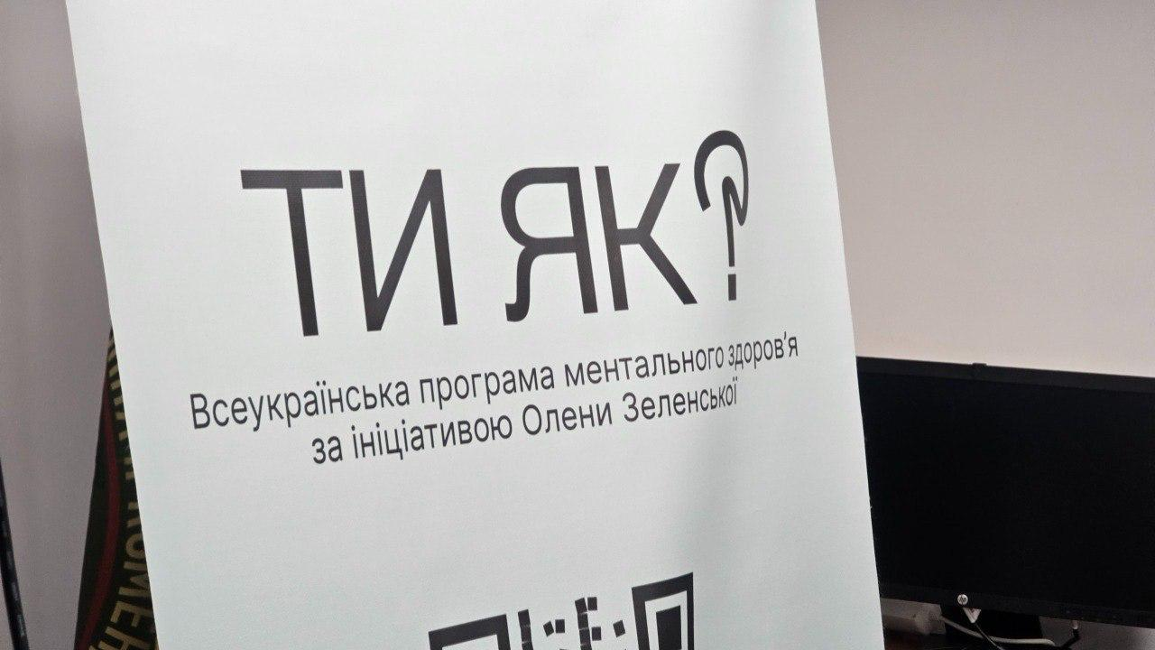 Білий інформаційний стенд із великим чорним логотипом програми «Ти як?» та підписом: «Всеукраїнська програма ментального здоров'я за ініціативою Олени Зеленської».