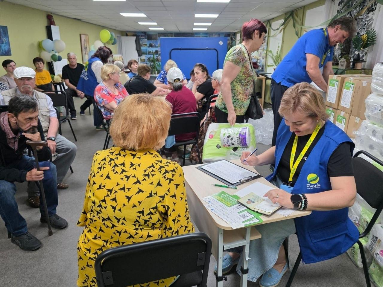 Процес реєстрації та видачі гуманітарної допомоги в приміщенні хабу. На передньому плані представниця благодійної організації «Рокада» у синьому жилеті вносить дані з паспорта відвідувачки. На задньому плані інша волонтерка складає коробки з допомогою, а група людей похилого віку чекає у черзі.