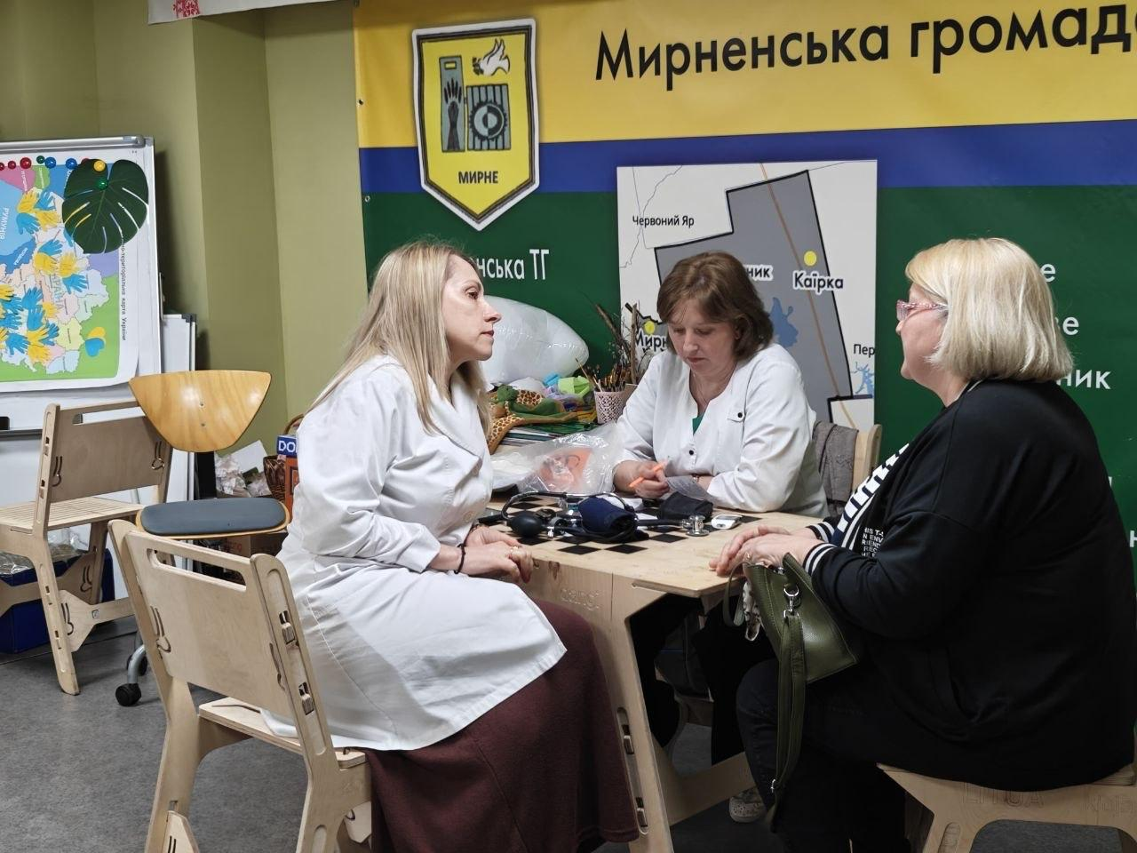 Дві медичні працівниці у білих халатах за столом консультують жінку на тлі великого синьо-жовто-зеленого банера з написом «Мирненська громада». На столі лежить тонометр для вимірювання тиску та медичні документи.