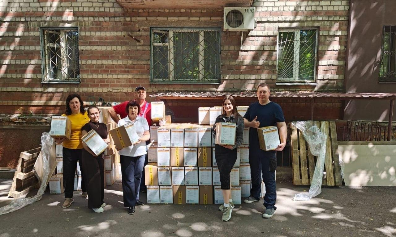 Група з шести людей стоїть на вулиці біля цегляного житлового будинку поруч із великою стопкою складених коробок із гуманітарною допомогою. Кожен тримає по одній коробці в руках, позуючи для фото.