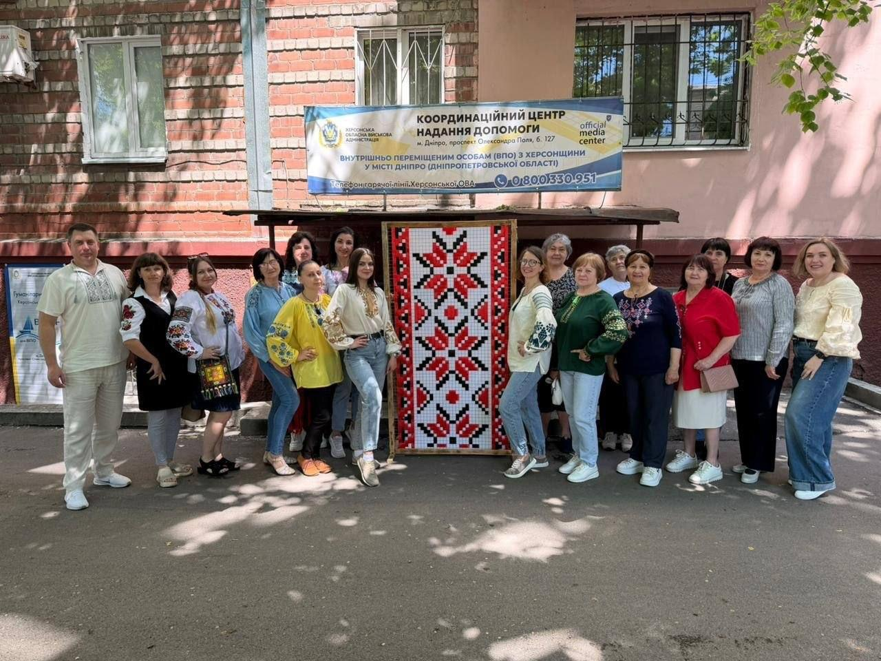 Велика група людей позує для спільного фото на вулиці біля будівлі Координаційного центру надання допомоги ВПО з Херсонщини у місті Дніпро. По центру встановлено велике вертикальне полотно з традиційним червоно-чорним українським орнаментом, а зверху розміщено інформаційний банер із контактами центру.